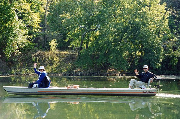 Wide shot of a man sitting at the end of a flat bottom float boat and a woman sitting  at the front of the boat, both waving their hands 