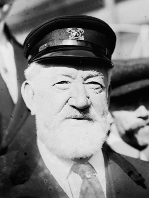 Up close black and white photo of an older white man with a white beard and a nautical hat squinting