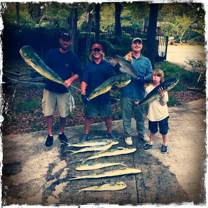 A group of three white men and one kid holding up mahi mahi fish with a few more displayed on the concrete ground.
