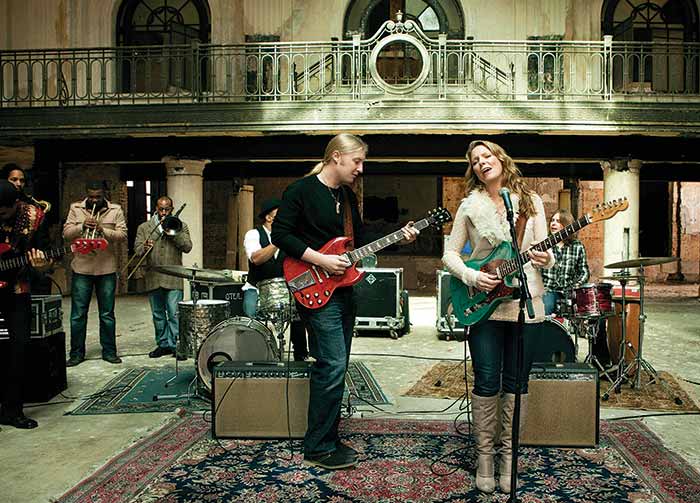 A white man and woman stand on an ornate black and red rug while singing and playing guitars in front of a band. 