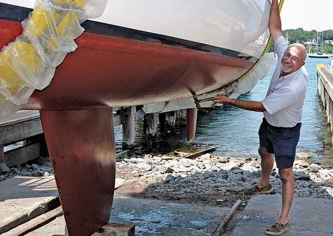 A man smiles and holds on to the side of a sailboat sitting on jacks on land