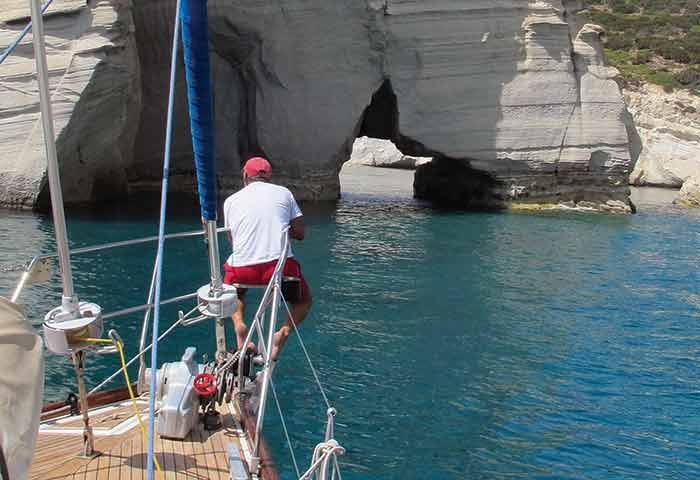 A man  in red shorts sits with his feet hanging off the front of a sailboat overlooking clear blue water