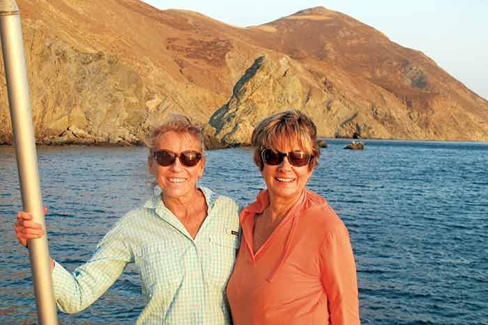 Two women in sunglasses smile while standing on a sailboat with the sea behind them
