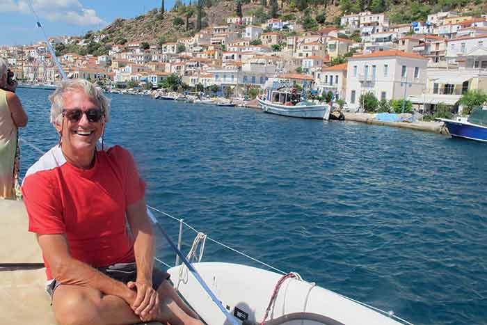 A man wearing sunglasses in a red shirt smiles at the camera from a sailboat