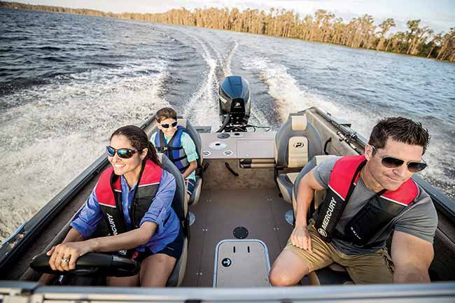 A women behind the helm of a fishing boat, a man sits to her left and a boy in the back.