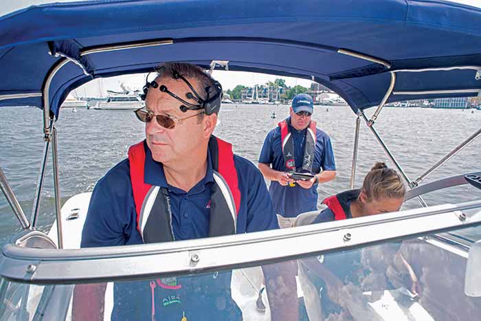 A man stands at the helm of the boat with black sensors attached to his head. Two others aboard the boat, one man, one woman with her head down.