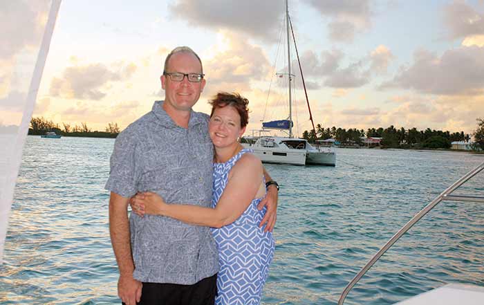 A man and a woman hug with a sunset and catamaran in the background