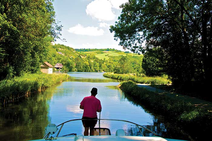Man standing on bow of a Le Boat self-drive barge meanders down the Canal du Nivernais, with a bike path along its banks