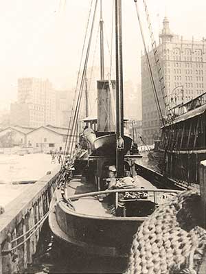 Black and white old photo of a sailboat docked with a large high-rise building in the background