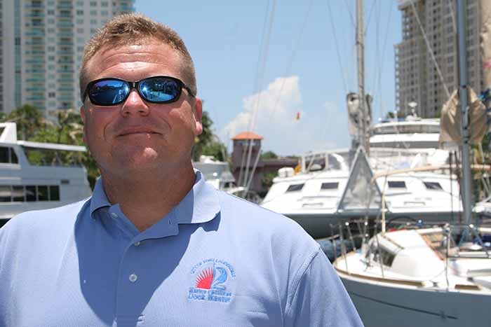 Man in light blue polo shirt and sunglasses standing infront of boats at a marina