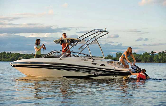 A wakeboard boat is stopped in the water as a woman assists a man in a lifejacket and his wakeboard back aboard
