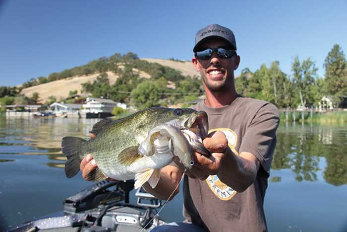 Man wearing a ball cap and sunglasses stands holding his big bass catch with Clear Lake in the background