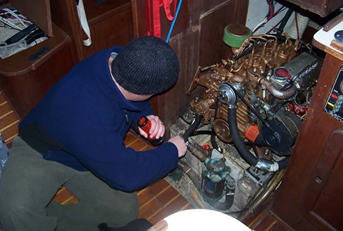 Man wearing a blue ling sleeve shirt and blue wool cap inspecting an inboard boat engine holding a flashlight