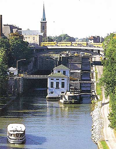 A boat navigates down the Erie Canal, a church steeple is visible in the background and a bridge containing several yellow school busses passes over the canal.