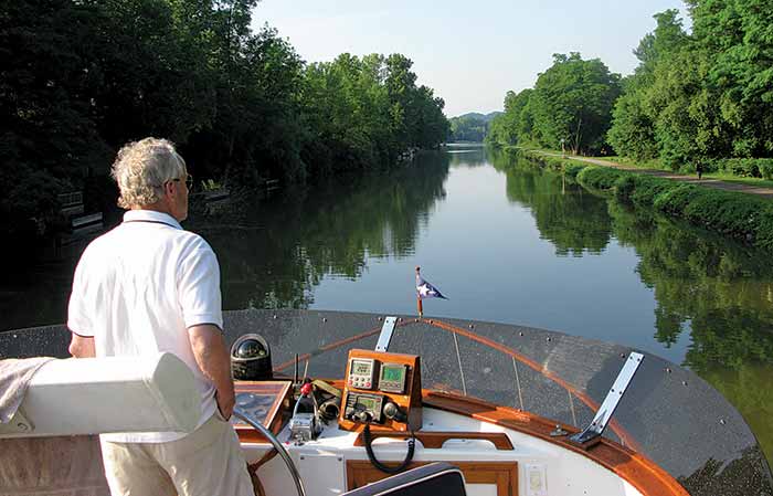 A man at the helm of a partially wooden boat overlooks calm water surrounded by green trees.