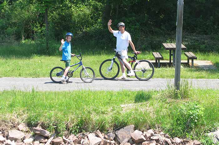 A man and a child ride their bikes through a park with many trees, waving hello