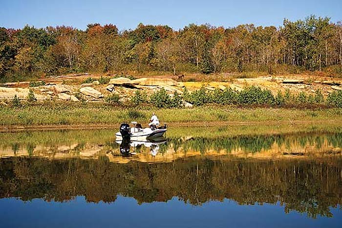 Small white boat with a man fishing in it on a clear blue lake with a rocky shoreline and tress in the background