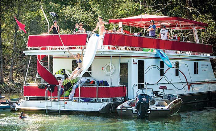 Red and white houseboat anchored along the shoreline with group of people enjoying swimming and lounging on the boat's deck