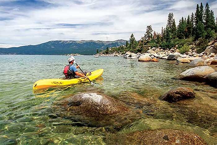 Man wearing a helmet, blue shirt and a red backpack paddling a bright yellow kayak over rock filled waters with trees and hills in the background