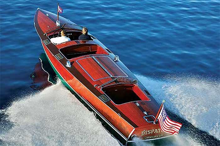 View from above of a couple cruising through the water in a sleak woooden powerboat with an American flag flying off the stern