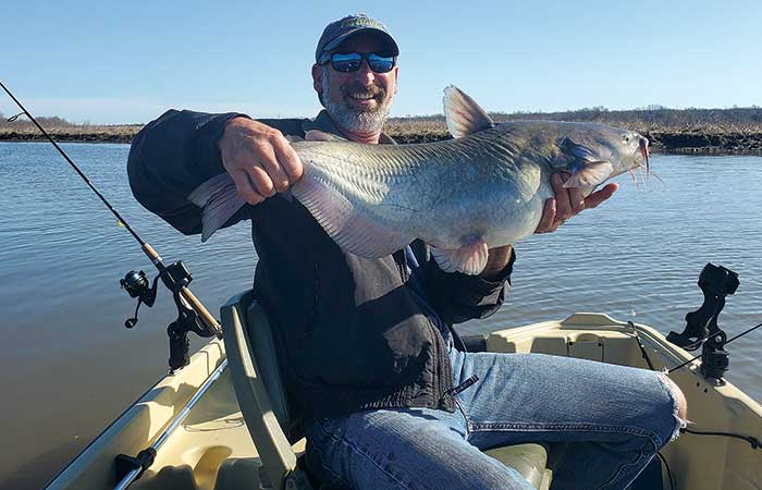 Man sitting in a small fishing boat holding up his large catfish catch