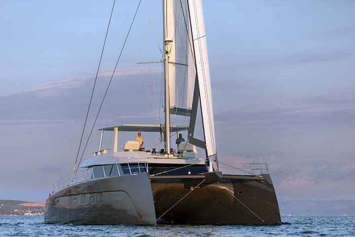 Two men at the helm of a CAT63 sailing yacht cruising along the water at sunset