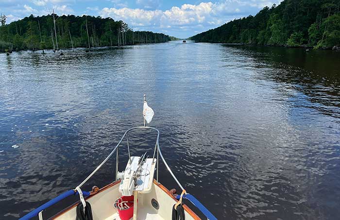 The bow of a boat with a small white falg cruising down a canal with trees on either side