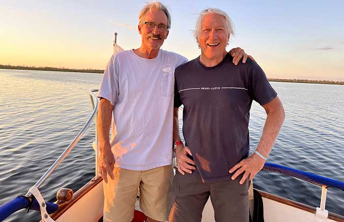 Two men standing on the bow of a boat on the water with one man with his arm arm the others shoulder as the sun sets in the background