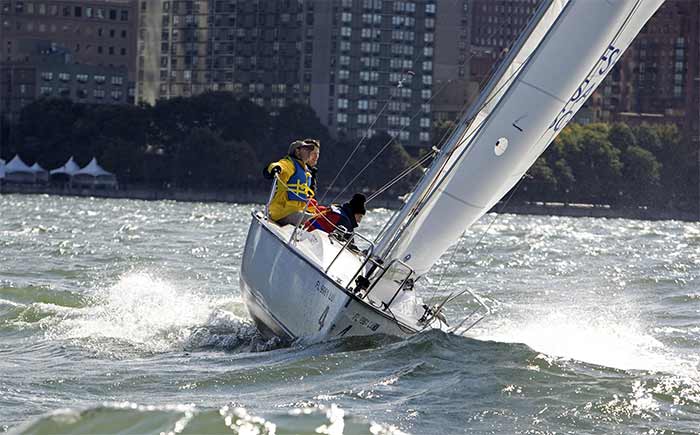 A couple sitting on a sailboat with a wave hitting the leeward bow and a cityscape in the background