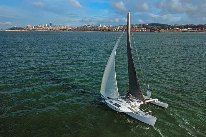 Man sailing a cataraman in open blue waters with a city skyline in the distance