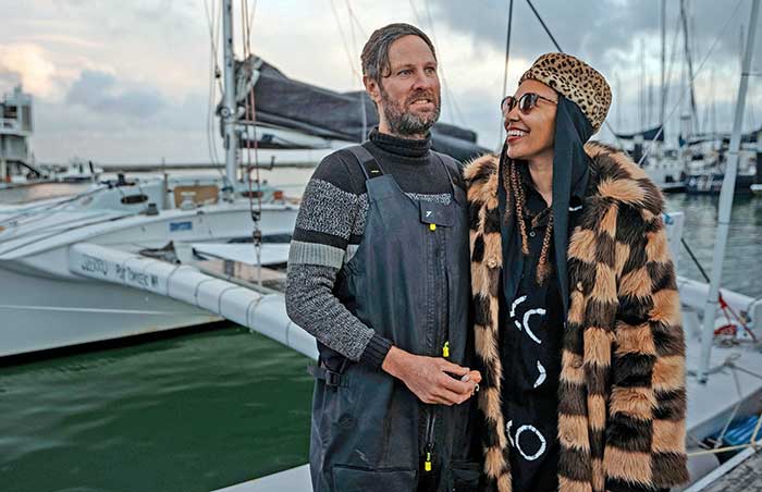Man wearing a gray and black stripped sweater and gray waders standing next to a women in wearing a brown and camel colored check jacket, leopard-print hat and sunglasses on the dock with a sailboat in the background