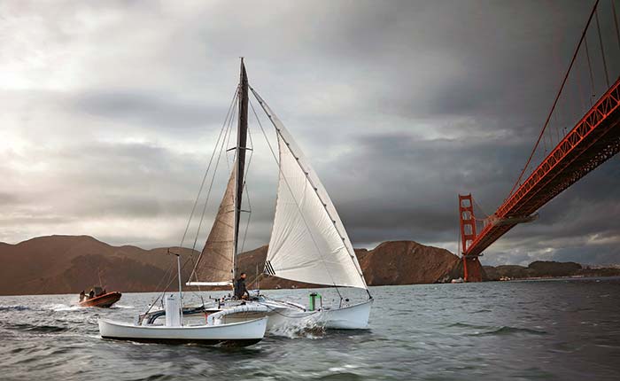 Man with beard and mustache wearing a blue jacket sailing a catamaran toward the Golden Gate Bridge in San Franciso wirh mountains in the background
