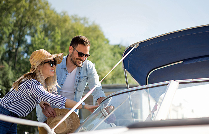 An adult female wearing sunglasses, a blue and white stripe shirt and tan hat next to an adult male wearing sunglasses and a denim shirt entering a boat.