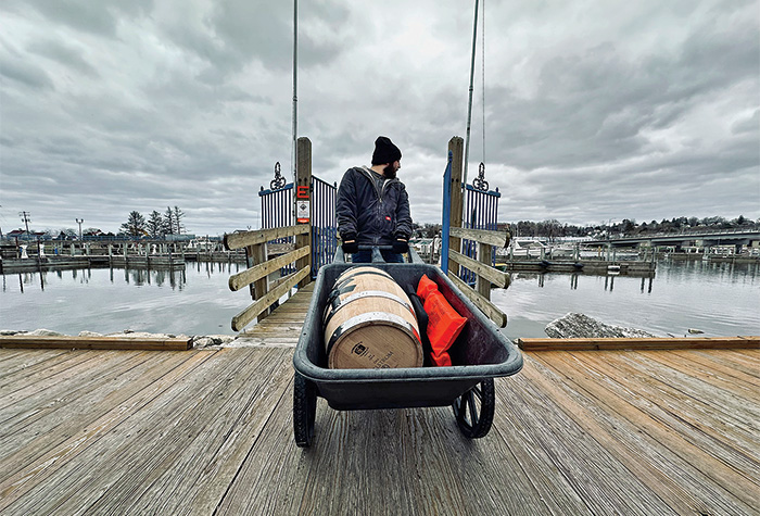 Adult male wearing a black stocking cap pulling a wheel barrel containing a whiskey barrel on a wooden dock. 