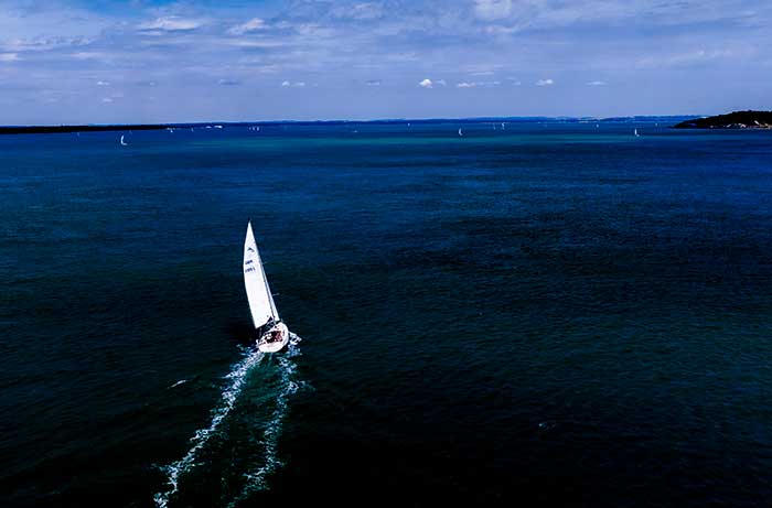 White sailboat sailing into open blue water with wake behind it and blue sky in the background