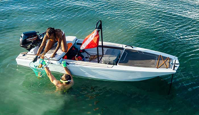 Man wearing a snorkle and mask holding onto the side of a small white boat putting a lobster into a net a woman kneeling on the side is holding