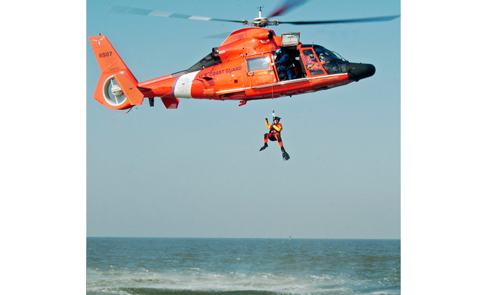 Large orange helicopter with rescue worker descending while hovering over open waters on a clear day. 