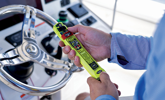 An adult holding a bright yellow rescue beacon over a silver steering wheel on a white vessel.