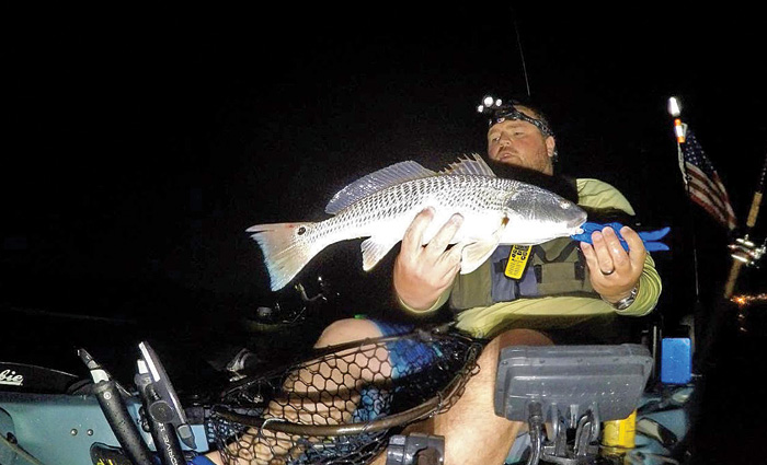 Adult male in the back of a fishing boat at night holding a caught fish.