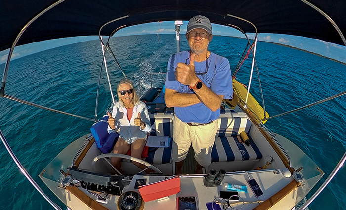 A senior male wearing a blue shirt and gray hat next to a female wearing sunglasses and a white shirt giving thumbs up on a boat at sea.