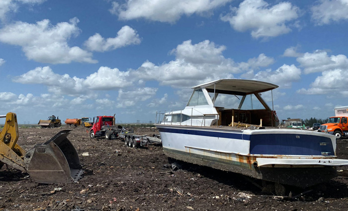 A rusted and old white and blue boat deserted in a junk yard towed by a red truck.