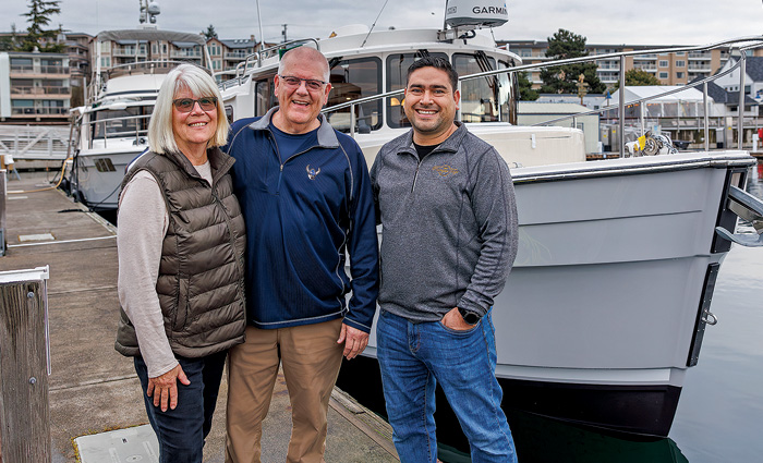 A woman and two men standing on a dock in front of a white boat during the day.