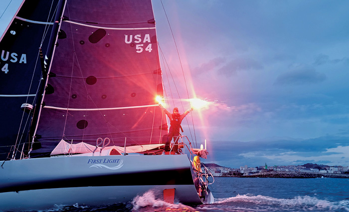 Adult holding two lit flares at the front of a sailboat during dusk on open waters.