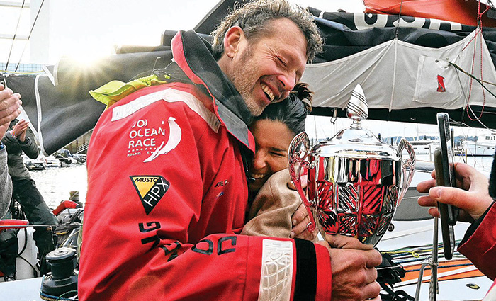 Adult male in a red rain jacket holding a trophy embracing a female with a tan blanket smiling.