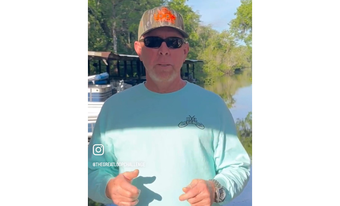 Adult male wearing a ballcap, black sunglasses and a light blue shirt standing on a dock in front of a lake.