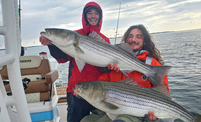 Two young adult males, one wearing a red rain coat and the other an orange coat, proudly displaying two large fish caught.