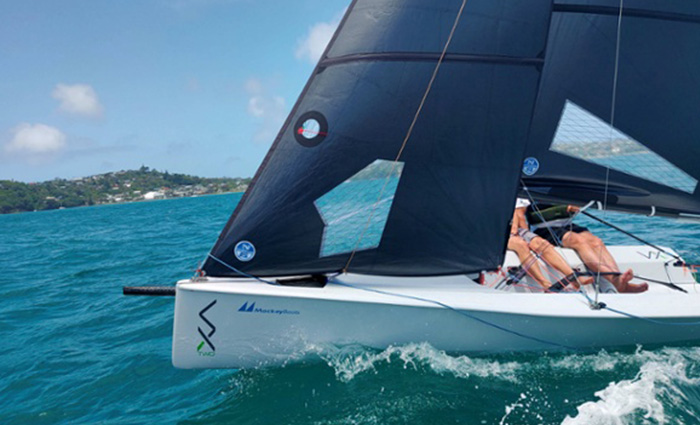 Two adults aboard a white sailboat with navy sails out on clear blue waters.