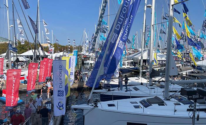 Numerous sailboats on display at a boat show with attendees walking alongside the dock on a clear day.