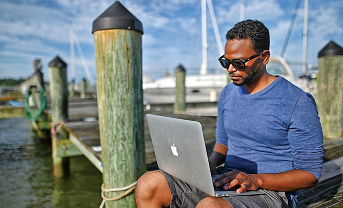 African-American adult male wearing a blue shirt, grey shorts and sunglasses sitting on a wooden dock and using a Mac laptop.
