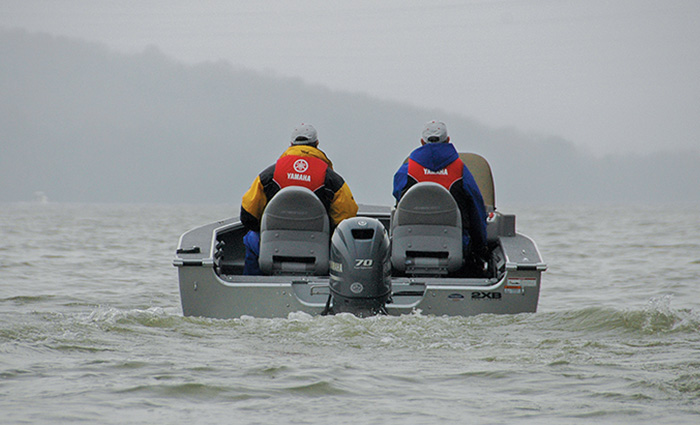 Two men on a small gray fishing boat during dense fog on open waters.